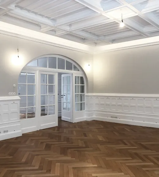 Sanierter Wohnraum der Stadtvilla R mit weißer Kassettendecke, Wandvertäfelung, Fischgrätparkett und Rundbogentür