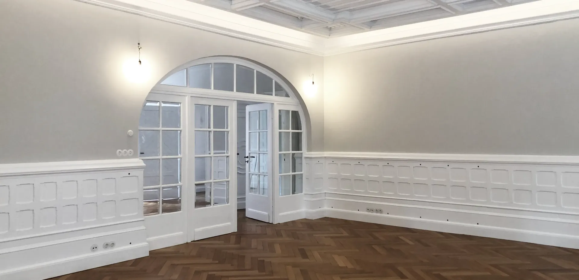 Sanierter Wohnraum der Stadtvilla R mit weißer Kassettendecke, Wandvertäfelung, Fischgrätparkett und Rundbogentür